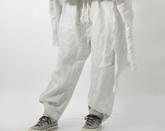 May include: A white linen jumpsuit with a loose fit and a drawstring waist. The jumpsuit has a long, flowing skirt and a short-sleeved top. The model is wearing the jumpsuit with a pair of black sneakers.