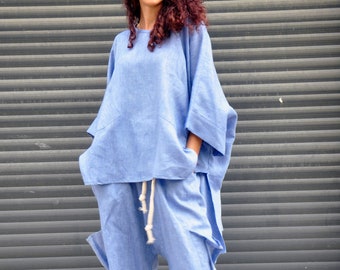 May include: A woman wearing a light blue linen pantsuit with a loose-fitting top and wide-leg pants. She is standing in front of a gray metal garage door and wearing pink sneakers.