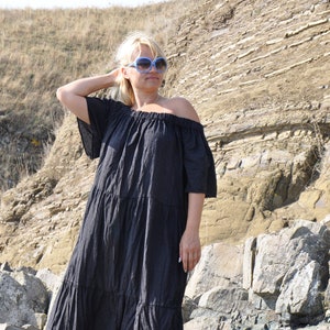 May include: A woman wearing a black off-the-shoulder maxi dress with a tiered skirt and blue sunglasses. She is standing on a rocky beach.