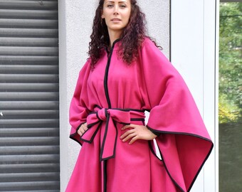 Könnte beinhalten: Ein asymmetrischer rosa Mantel mit schwarzem Besatz und einem schwarzen Gürtel. Der Mantel hat eine lange, fließende Silhouette und ist mit einem Gürtel in der Taille gebunden.