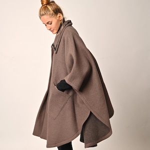 Op de afbeelding: Een bruine capejas met een kraag en twee voorzakken. De jas is knielang en heeft een split aan de achterkant. De persoon draagt ​​zwarte leggings en zwarte laarzen.