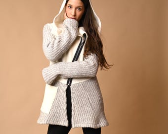 May include: A hooded, textured knit coat in gray and cream, featuring a front zipper and a unique asymmetrical design. The coat is paired with black pants and black boots, creating a stylish, layered look.