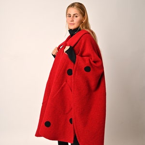 Pode incluir: Um poncho de lã vermelho com bolinhas pretas e gola alta preta. O poncho tem um caimento solto e está sendo usado por uma pessoa com cabelo loiro comprido.