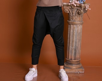 May include: A woman wearing a brown long-sleeved shirt with a black and white design and black pants with a loose fit. She is standing in front of a brown background with a decorative column behind her.