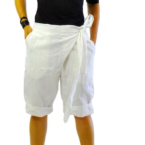 May include: A person wearing a black short-sleeved top, white wrap shorts, and black platform sandals with sunglasses. The shorts have a tie closure. The background is white.
