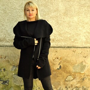 K&ouml;nnte beinhalten: Eine Frau tr&auml;gt einen schwarzen Langarmmantel mit gro&szlig;em Kragen und Rei&szlig;verschluss. Sie tr&auml;gt eine schwarze Leggings und schwarze Stiefel.
