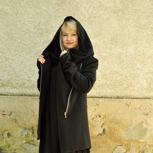 K&ouml;nnte beinhalten: Eine schwarze Kapuzenjacke mit einem langen, asymmetrischen Saum. Die Jacke hat einen Rei&szlig;verschluss und einen hohen Kragen.