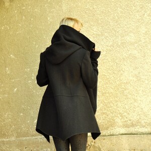 K&ouml;nnte beinhalten: Ein schwarzer Kapuzenmantel mit einem langen, asymmetrischen Saum. Der Mantel wird von einer Person mit blonden Haaren getragen, die auf einer Steintreppe steht.