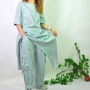 A woman in a light green linen tunic and cargo pants set, with sneakers, stands with one hand holding up the tunic's hem.