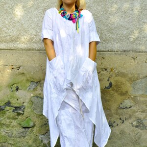 A woman in a white linen tunic and pants set, with a colorful fabric necklace, stands with hands in the tunic's pockets.