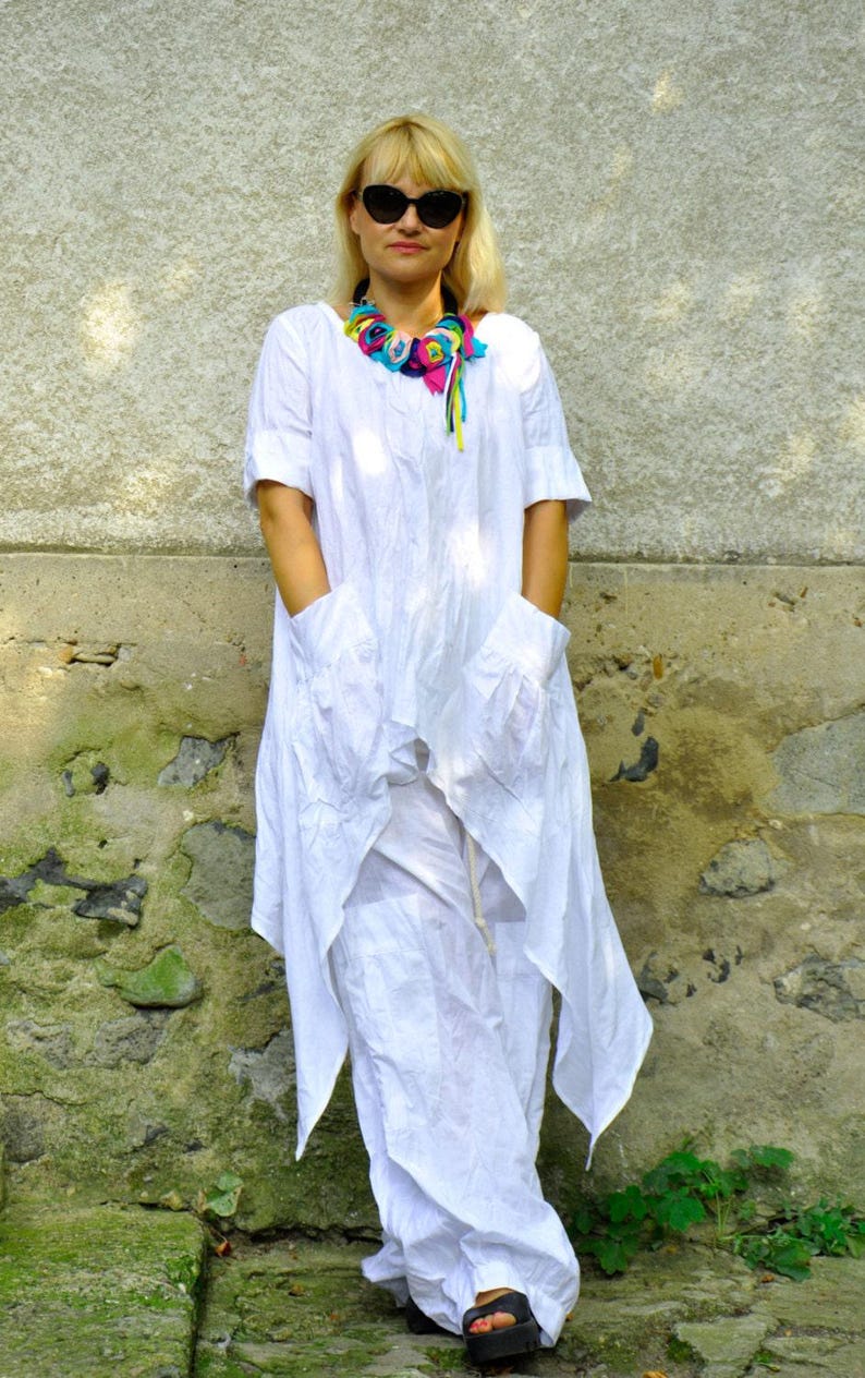 A woman in a white linen tunic and pants set, with a colorful fabric necklace, stands with hands in the tunic's pockets.