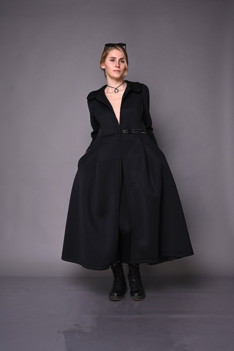 May include: A black long-sleeved dress with a deep V-neckline and a belt at the waist. The dress has a flowing skirt and is worn with black boots.