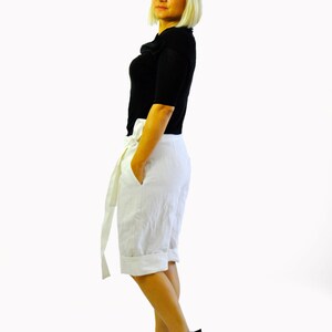 May include: A person wearing a black top, white shorts with a tie, and black platform sandals. They are also wearing black sunglasses. The outfit is set against a white background.
