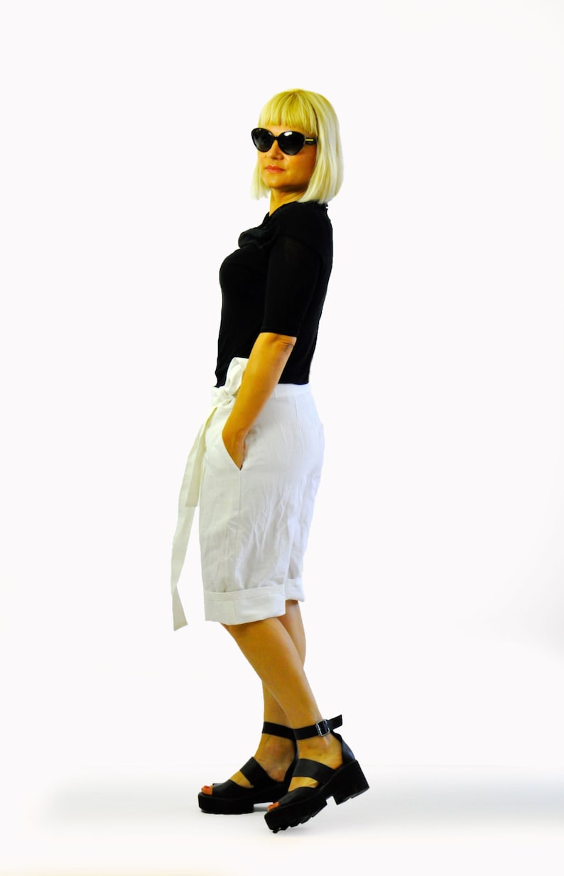 May include: A person wearing a black top, white shorts with a tie, and black platform sandals. They are also wearing black sunglasses. The outfit is set against a white background.