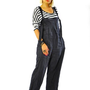 May include: Dark gray linen overalls with adjustable straps and wide legs. The overalls are paired with a black and white striped long-sleeve shirt and black platform sandals. The model is wearing sunglasses.
