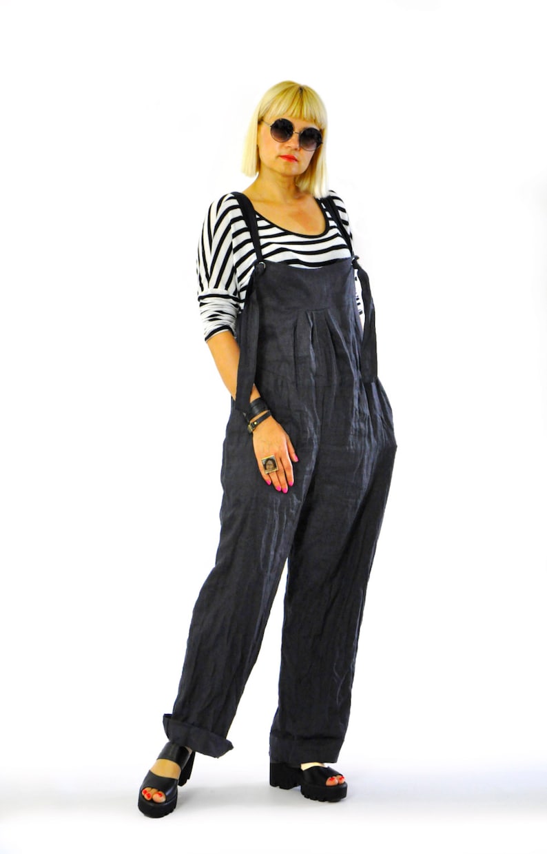 May include: Dark gray linen overalls with adjustable straps and wide legs. The overalls are paired with a black and white striped long-sleeve shirt and black platform sandals. The model is wearing sunglasses.