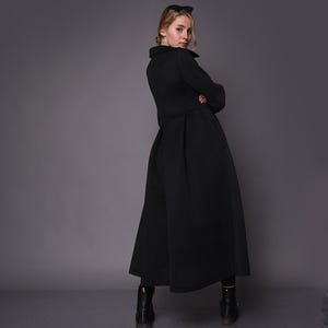 A model seen from the back, wearing a long black coat with a structured pleated waist and dark boots.