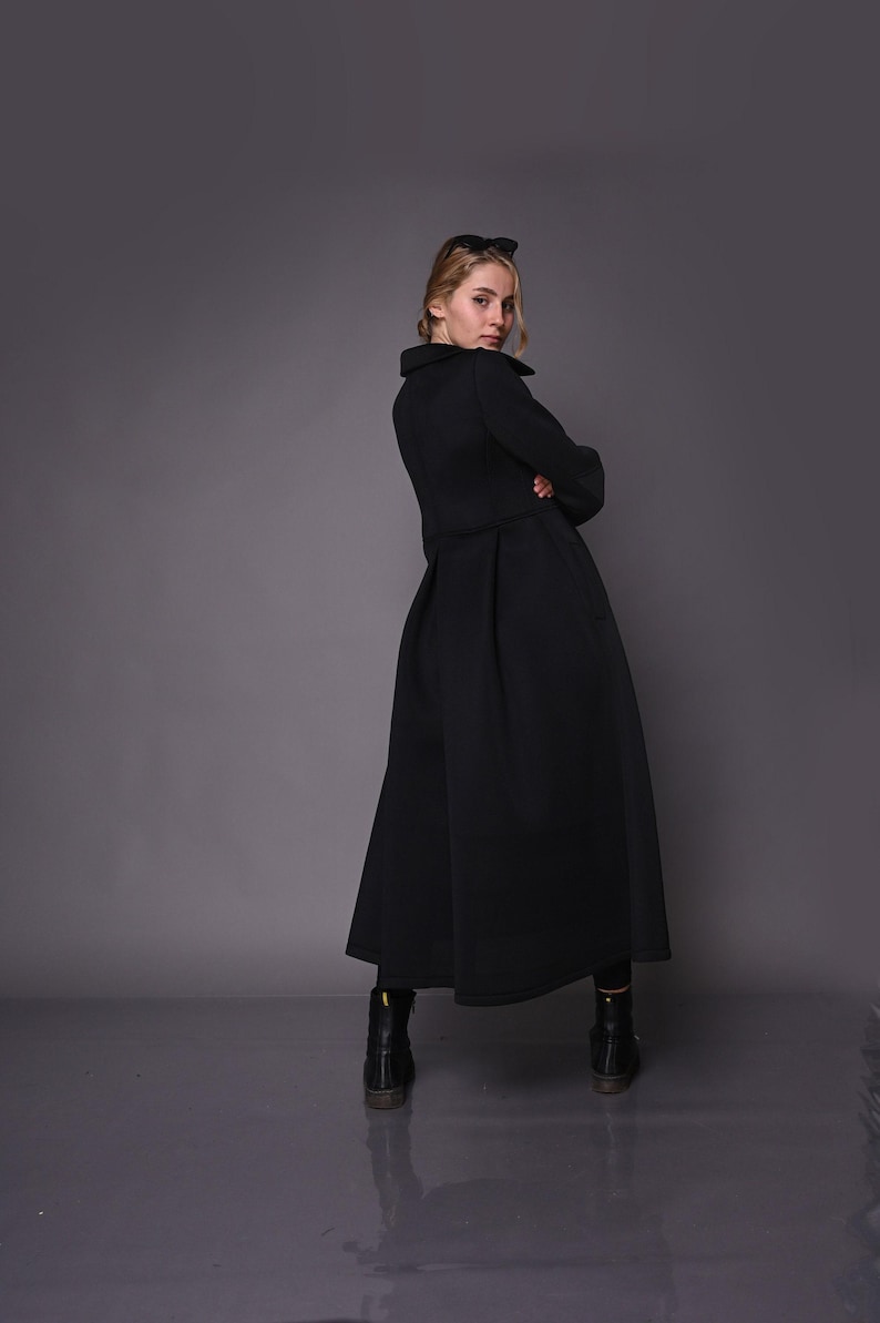 A model seen from the back, wearing a long black coat with a structured pleated waist and dark boots.