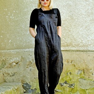 May include: A woman wearing a black jumpsuit with a black short-sleeved top underneath. The jumpsuit has large pockets and adjustable straps. She is wearing white sunglasses and black shoes. The background is a textured, light-colored wall.