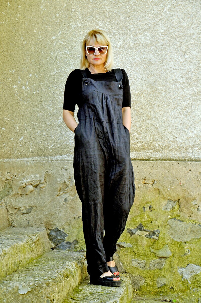 May include: A woman wearing a black jumpsuit with a black short-sleeved top underneath. The jumpsuit has large pockets and adjustable straps. She is wearing white sunglasses and black shoes. The background is a textured, light-colored wall.