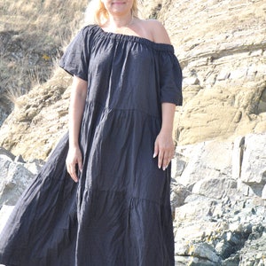 May include: A woman wearing a black off-the-shoulder maxi dress with a tiered skirt and blue sunglasses. She is standing on a rocky beach.