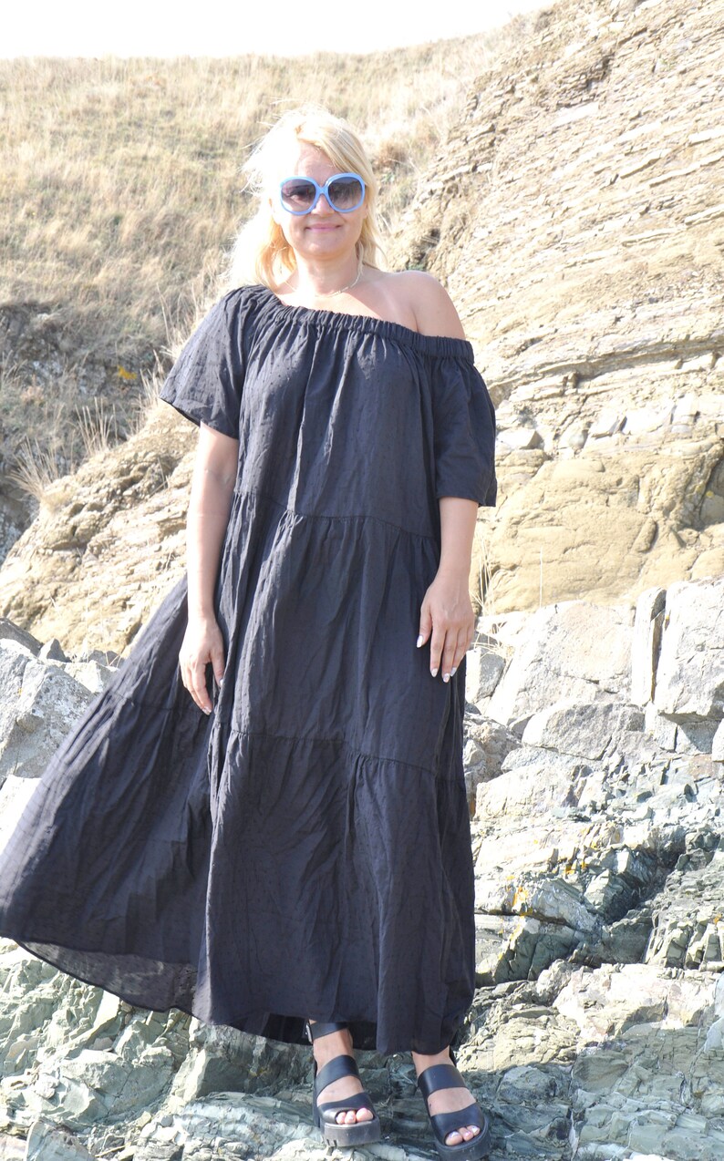 May include: A woman wearing a black off-the-shoulder maxi dress with a tiered skirt and blue sunglasses. She is standing on a rocky beach.