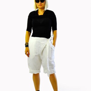 May include: A person wearing a black short-sleeved top and white wrap shorts. The outfit is paired with black platform sandals and sunglasses. The shorts have a tie closure at the waist. The background is white.