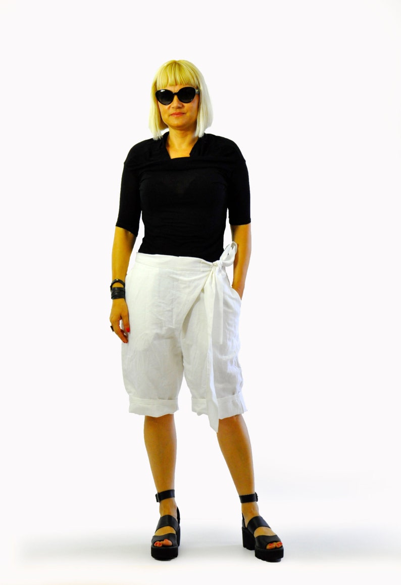 May include: A person wearing a black short-sleeved top and white wrap shorts. The outfit is paired with black platform sandals and sunglasses. The shorts have a tie closure at the waist. The background is white.