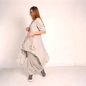 A woman with long blonde hair looks down, wearing a light gray linen tunic with large front pockets and an asymmetrical hem, paired with matching wide-leg pants and gray sneakers.