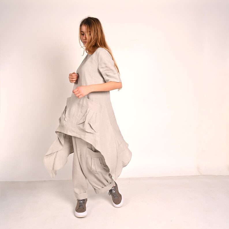 A woman with long blonde hair looks down, wearing a light gray linen tunic with large front pockets and an asymmetrical hem, paired with matching wide-leg pants and gray sneakers.