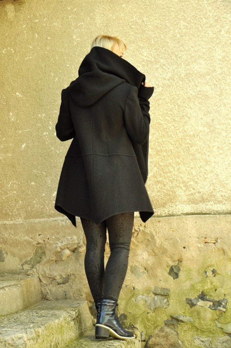 K&ouml;nnte beinhalten: Ein schwarzer Kapuzenmantel mit einem langen, asymmetrischen Saum. Der Mantel wird von einer Person mit blonden Haaren getragen, die auf einer Steintreppe steht.