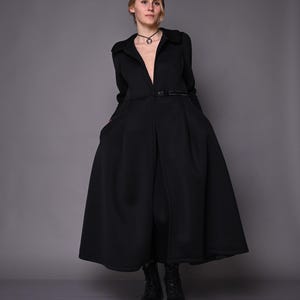 A model posing in a long black winter coat with a belted waist and flared skirt, standing on a grey floor.