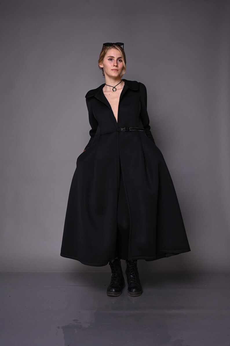A model posing in a long black winter coat with a belted waist and flared skirt, standing on a grey floor.