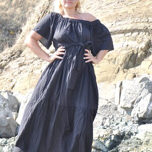 May include: A woman wearing a black off-the-shoulder maxi dress with a tie waist and blue sunglasses. She is standing on a rocky beach.