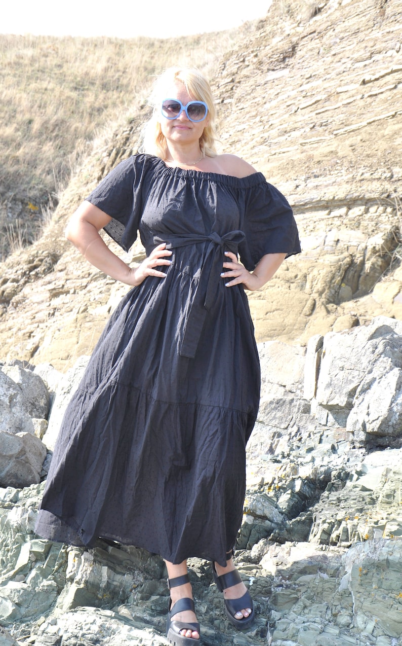 May include: A woman wearing a black off-the-shoulder maxi dress with a tie waist and blue sunglasses. She is standing on a rocky beach.