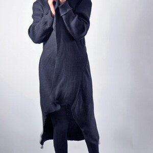 A front view of a woman holding the collar of a long black hooded cardigan closed, wearing white boots.