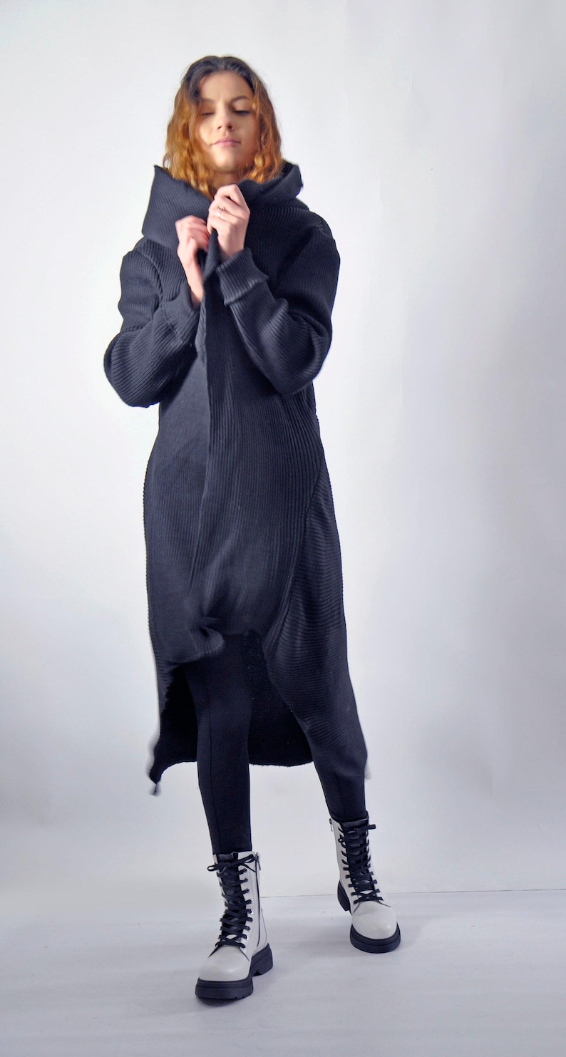 A front view of a woman holding the collar of a long black hooded cardigan closed, wearing white boots.