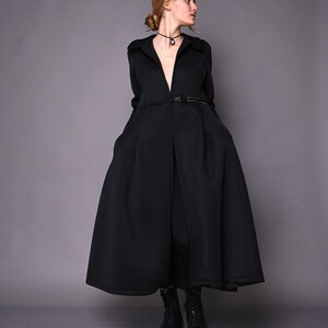 A model posing in a long black princess-style coat with her hands in the pockets, viewed from the front-right side.