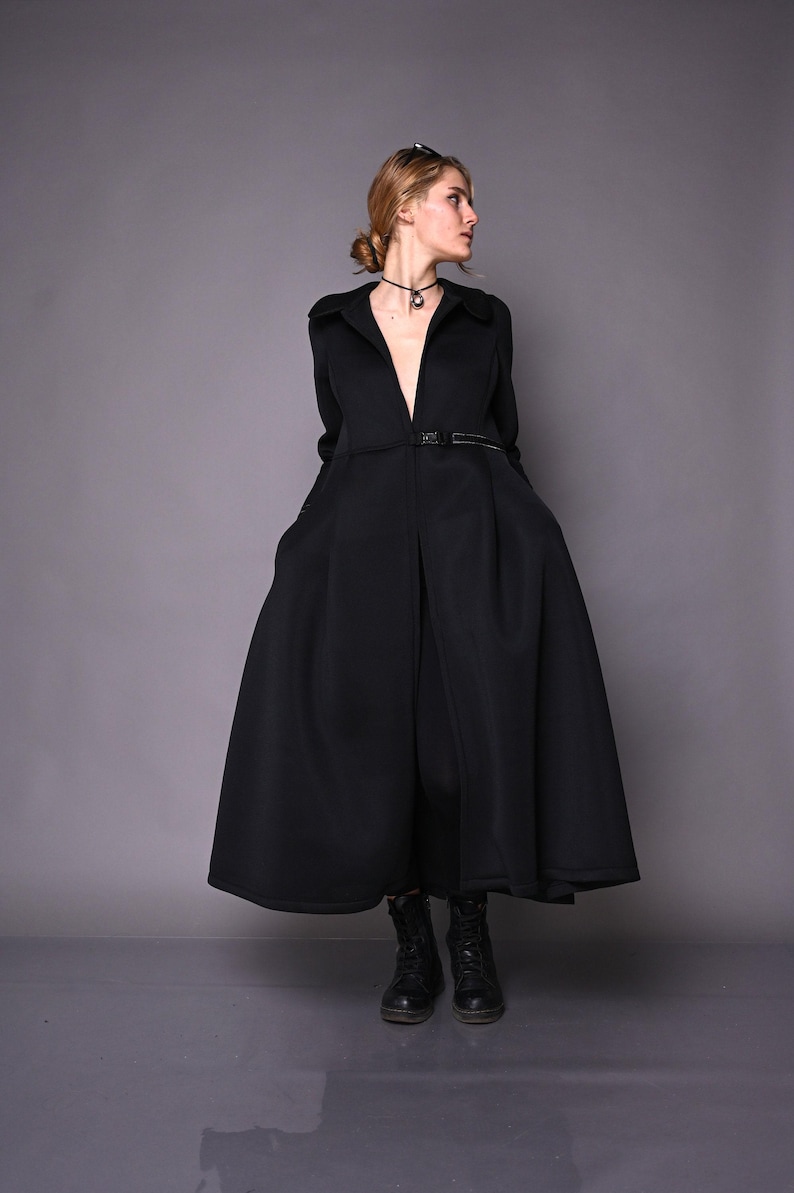A model posing in a long black princess-style coat with her hands in the pockets, viewed from the front-right side.