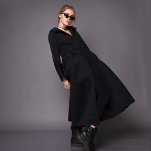 May include: A black long-sleeved coat with a high collar and a flared skirt. The coat has a button closure and a belt. The model is wearing black sunglasses and black boots.