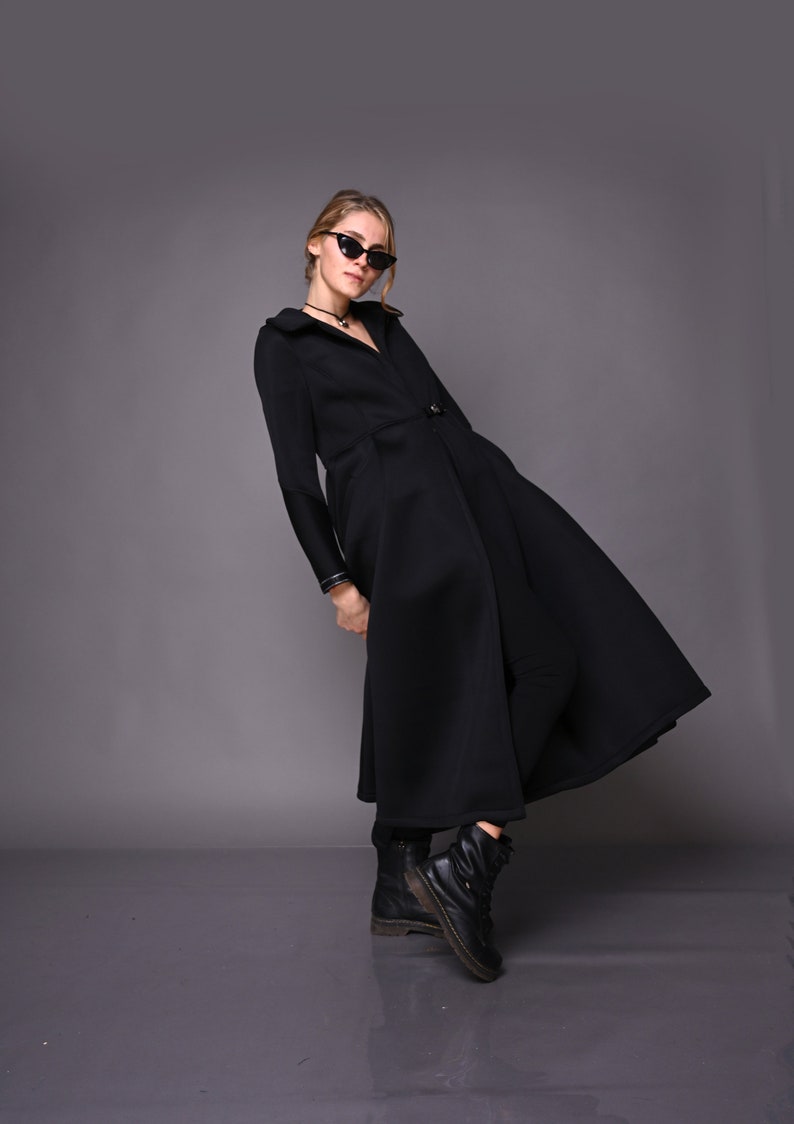 May include: A black long-sleeved coat with a high collar and a flared skirt. The coat has a button closure and a belt. The model is wearing black sunglasses and black boots.