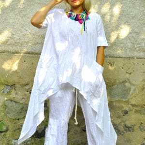 A woman in a white linen tunic and pants set, with a colorful fabric necklace, stands with one hand on her hip and the other holding sunglasses.