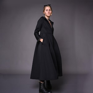 A model standing at an angle in a long black winter coat, showcasing the fitted bodice and flared maxi hem.