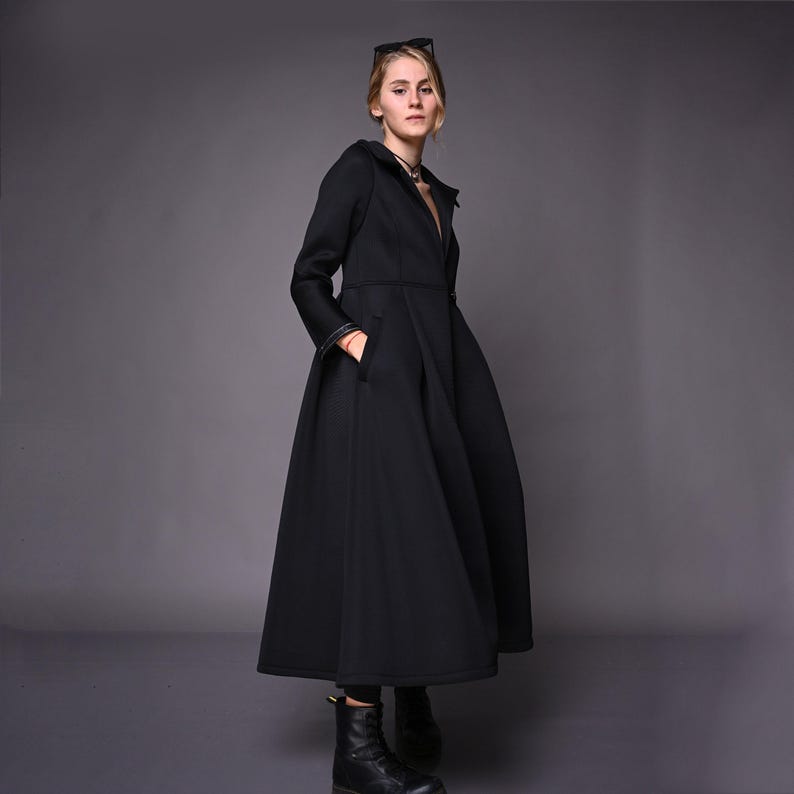 A model standing at an angle in a long black winter coat, showcasing the fitted bodice and flared maxi hem.