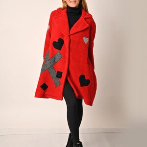 May include: A red fleece coat with black and grey heart-shaped patches. The coat has a collar and a button closure. The model is wearing black leggings and black boots.