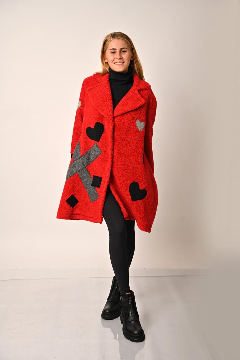 May include: A red fleece coat with black and grey heart-shaped patches. The coat has a collar and a button closure. The model is wearing black leggings and black boots.