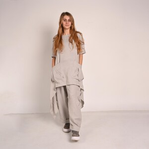 A woman with long blonde hair stands facing forward, wearing a light gray linen tunic with an asymmetrical hem and wide-leg light gray linen pants with large pockets.