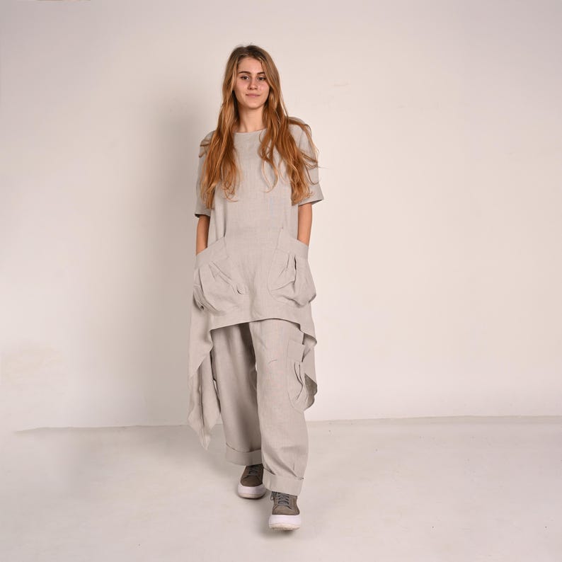 A woman with long blonde hair stands facing forward, wearing a light gray linen tunic with an asymmetrical hem and wide-leg light gray linen pants with large pockets.