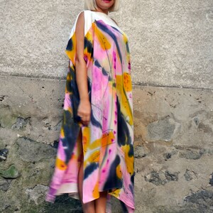 Puede incluir: Una mujer con un vestido maxi de estampado abstracto blanco y rosa, un top blanco y gafas de sol blancas. Est&aacute; de pie frente a una pared de piedra.