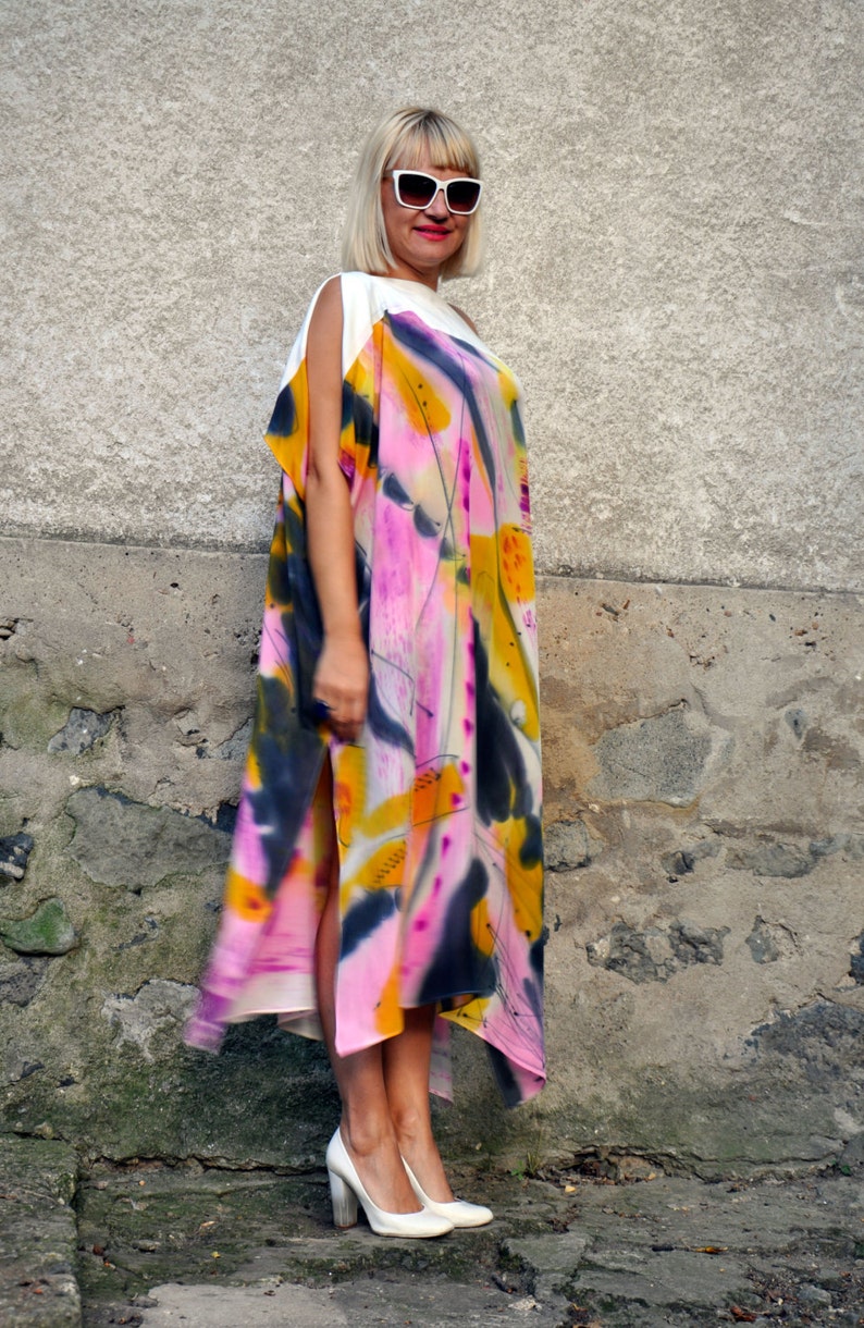 Puede incluir: Una mujer con un vestido maxi de estampado abstracto blanco y rosa, un top blanco y gafas de sol blancas. Est&aacute; de pie frente a una pared de piedra.